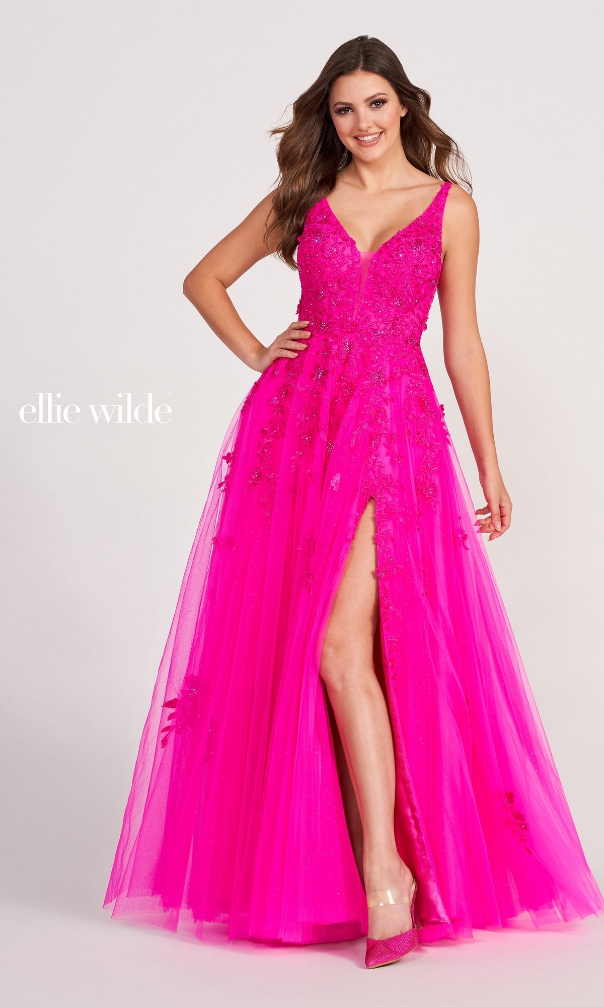 A woman poses confidently in the Ellie Wilde Side-Slit Long Prom Ball Gown EW34103, featuring a bright pink hue, sleeveless design, floral lace details, deep V-neckline, and a high slit.